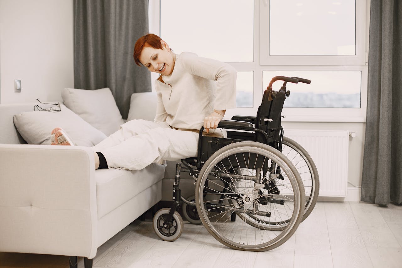 A woman with dyed short hair smiles while transitioning from a wheelchair to a couch.