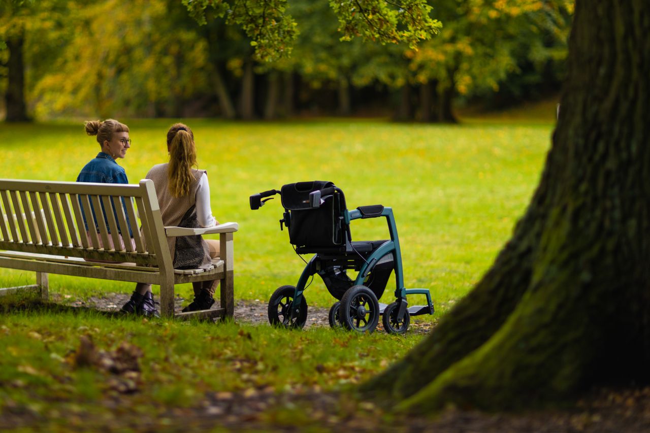 services-01 Two women sitting on a bench in a park beside a wheelchair, enjoying a peaceful day outdoors.
