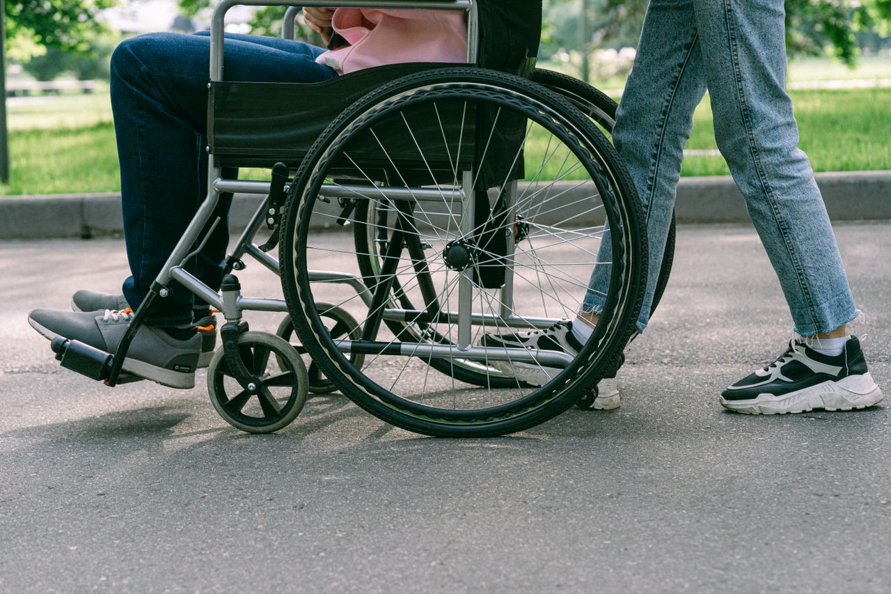 about-01 Close-up view of a person in a wheelchair being assisted on a sunny urban pavement.